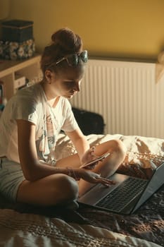 Young woman using portable computer and mobile phone, sitting on a bed, learning online at home. Candid people, real moments, authentic situations