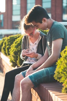 Couple of friends, teenage girl and boy,  having fun together with smartphones,  sitting in center of town, spending time together