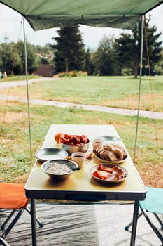 Breakfast prepared during summer vacation on camping. Bread, cottage cheese, cold meat, tomatoes, fruits and coffee cups on table. Close up of outdoor table setting set under tent