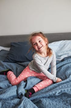 Little happy playful kid girl having fun making silly funny faces jumping on bed in bedroom in the morning at home