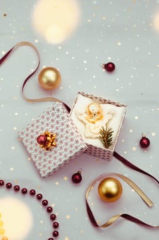 Wrapping Christmas gift into box. Decorating using red ribbon, red Christmas balls and pine twigs. Pink fabric in the background. Blurred Christmas lights. Flat top-down composition. Figure of angel made of earthen with love inscription as Christmas decoration
