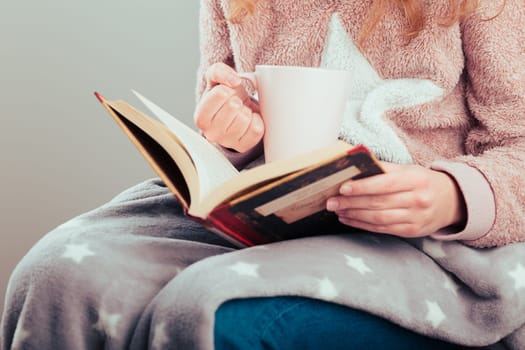 Girl enjoying the reading a book and drinking coffee at home. Young woman sitting on a chair, wrapped in blanket, holding book, relaxing at home. Portrait orientation. Front view