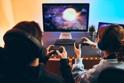 Friends playing video game at home. Gamers holding gamepads sitting at front of screen. Streamers girl and boy playing online in dark room lit by neon lights. Competition and having fun