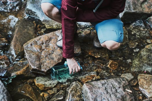 Woman taking pure water to bottle from mountain stream during trekking in mountains. Hiker crouching on rocks, filling bottle up with cold mountain water. Enjoying the outdoors in the summer trip vacation