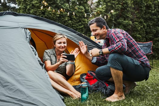 Couple having video call with friends using smartphone while sitting in tent at camping. People relaxing in tent during summer vacation. Actively spending vacations outdoors close to nature. Concept of camp life