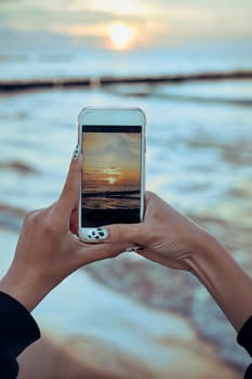 Young woman taking photos of sunset over sea using smartphone during summer trip. Rear view of girl holding smartphone and taking picture