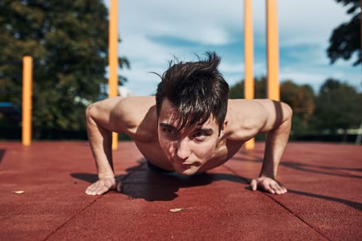 Young shirtless man bodybuilder doing push-ups on a red rubber ground during his workout in a modern calisthenics street workout park