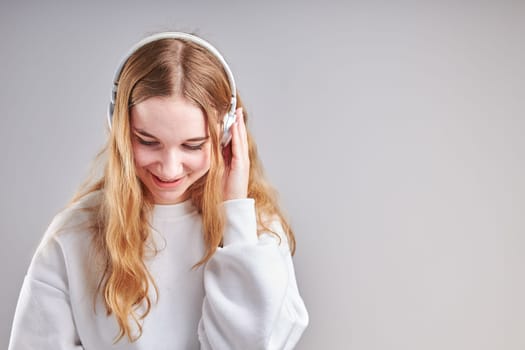 Young woman girl listening to music streaming content having fun watching video enjoying video chat talking with friends making gestures faces using smartphone earphones headphones standing over plain grey background