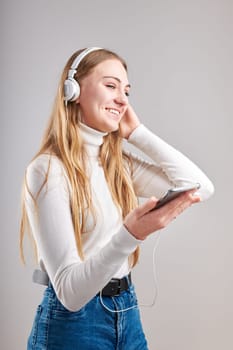 Young woman girl listening to music streaming content having fun watching video enjoying video chat talking with friends making gestures faces using smartphone earphones headphones standing over plain grey background
