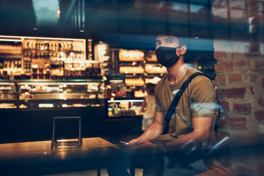 Young man sitting in coffee shop at store front in the city center in the evening, wearing the face mask to avoid virus infection and to prevent the spread of disease in time of coronavirus