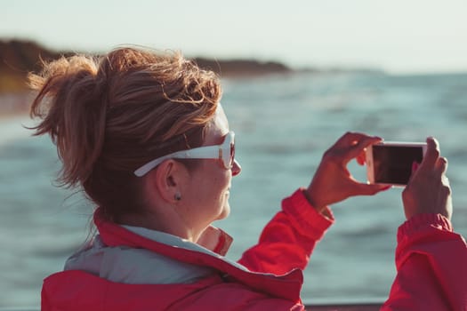 Young woman taking a photos using phone, looking at screen, standing outdoors, she is backlighted by sunlight with plain sky and sea in the background