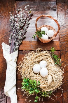Easter composition of willow twigs with catkins, boxwood, eggs in a easter basket and straw nest, set on old wooden table. Catkins are wrapped up in paper. Inside top shot. Vertical image.