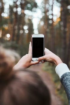 Young woman taking photos using smartphone during walk in a forest on summer vacation trip. Back view of girl holding smartphone