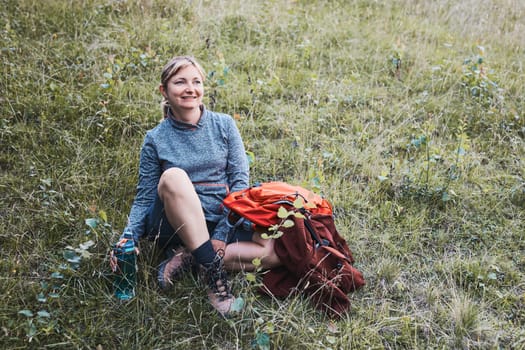 Smiling happy woman with backpack resting on grass during a hike in the mountains, actively spending summer vacation