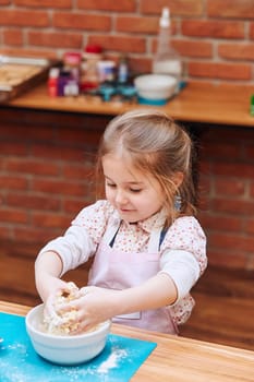 Little girl kneading the dough for baking the cake. Kid taking part in baking workshop. Baking classes for children,  aspiring little chefs. Learning to cook. Combining and stirring prepared ingredients. Real people, authentic situations