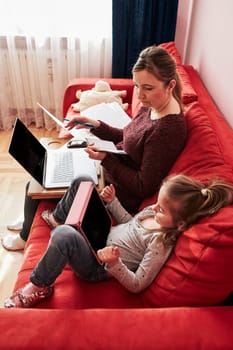 Woman mother working doing her job remotely during video chat call stream online course webinar on laptop from home during COVID-19 quarantine while her daughter watching video on tablet. Woman sitting on bed in front of computer looking at screen
