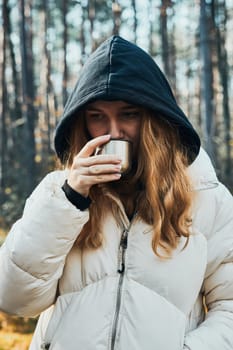 Woman in a hood having break during autumn trip holding cup with hot drink from thermos flask on autumn cold day. Active girl wandering in a forest actively spending time