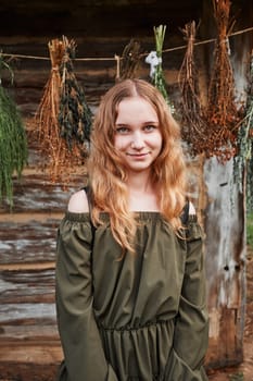 Portrait of young woman  standing in front of old cottage and hung some withered species of herbs. Girl is wearing green dress, looking at camera