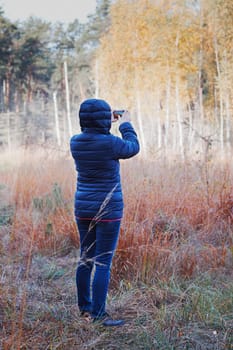 Woman taking photo of forest using camera in mobile phone. Forest in autumn season. Colorful foliage on trees lit by morning sunlight. Natural nature forest landscape in autumn warm sunlight day