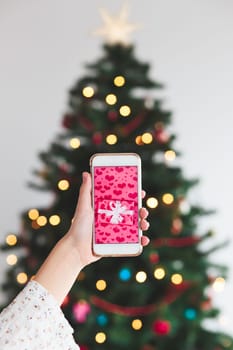 Girl holding the smartphone with wrapped gift on the screen and christmas tree in the background