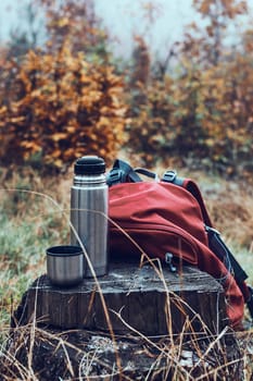 Close shot of thermos flask and backpack put by tree trunk while break during autumn trip on autumn cold day