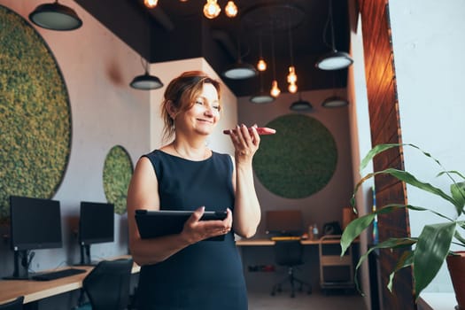 Businesswoman having business call working on tablet in office. Mature busy woman using touch pad computer standing by window in modern interior. Female manager focused on work holding digital device. Using technology