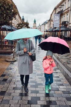 Mother and her little daughter holding the pink and blue umbrellas walking in a downtown on rainy gloomy autumn day