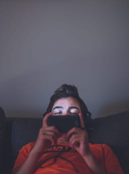 Boy using smart phones, listening to music, playing the games sitting on a couch at home