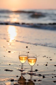 Two wine glasses with white wine standing on sand on beach in the foreground. Blurred couple standing in the sea in the background