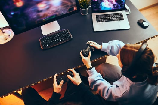 Friends playing video game at home. Gamers holding gamepads sitting at front of screen. Streamers girl and boy playing online in dark room lit by neon lights. Competition and having fun