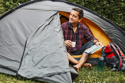 Man relaxing in tent at camping during summer vacation. Man holding map while planning next trip. Actively spending vacations outdoors close to nature. Concept of camp life