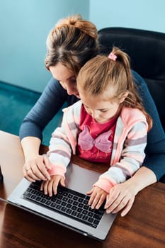 Little girl preschooler learning online solving puzzles playing educational games typing letters listetning to music and sounds with help of her mother using laptop computer and sitting at desk at home
