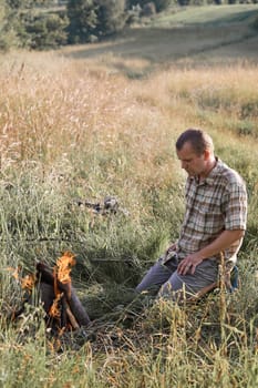 Man sitting in a grass beside a campfire on a meadow. Candid people, real moments, authentic situations
