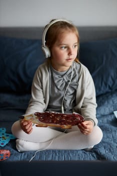Little girl preschooler learning online showing her works drawings done at home. Child learning watching lesson remotely, talking with tutoress on video call from home during quarantine. Kid using laptop, headphones sitting on bed