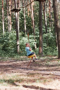 Happy boy riding on the zip line in rope park in forest while spending summer vacation. Real people, authentic situations