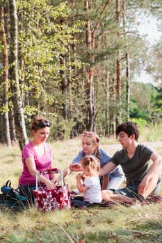 Family spending vacation time together having a snacks on a picnic sitting on blanket on grass in forest on sunny day in the summertime