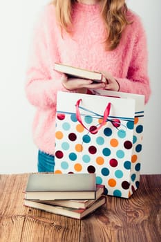 Young girl putting the books into paper bag in bookstore. A few books on a wooden table. Teenager girl wearing pink sweater and blue jeans. Vertical photo