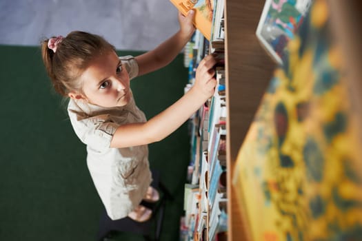 Sttudent looking at top bookshelf in school library. Smart girl selecting literature for reading. Books on shelves in bookstore. Learning from books. Back to school. Elementary education. Benefits of everyday reading. Curious child