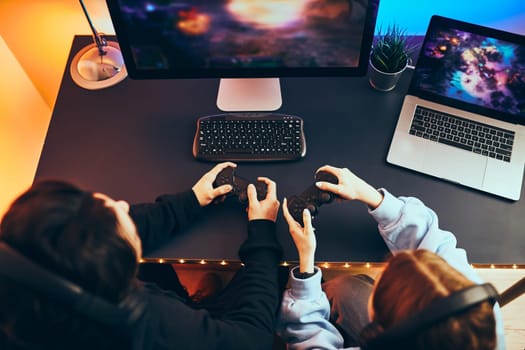 Friends playing video game at home. Gamers holding gamepads sitting at front of screen. Streamers girl and boy playing online in dark room lit by neon lights. Competition and having fun