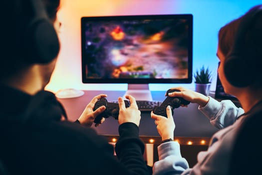 Friends playing video game at home. Gamers holding gamepads sitting at front of screen. Streamers girl and boy playing online in dark room lit by neon lights. Competition and having fun