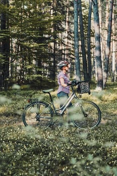 Active woman spending free vacation time on a bicycle trip in a forest on sunny summer day. Woman wearing bicycle helmet and gloves holding bike with basket standing behind the blurred tree leaves