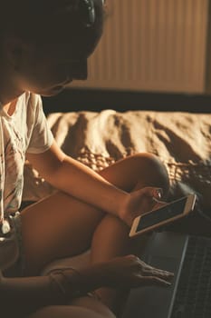 Young woman using portable computer and mobile phone, sitting on a bed, learning online at home. Candid people, real moments, authentic situations