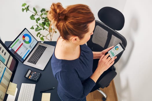 Businesswoman working with financial data on charts and tables on smartphone and computer. Woman entrepreneur standing by desk in office