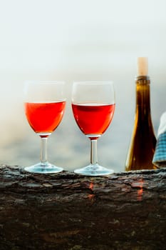 Two wine glasses with red wine standing on tree trunk, on beach, beside bottle of wine