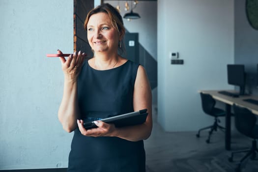 Businesswoman having business call working on tablet in office. Mature busy woman using touch pad computer standing by window in modern interior. Female manager focused on work holding digital device. Using technology