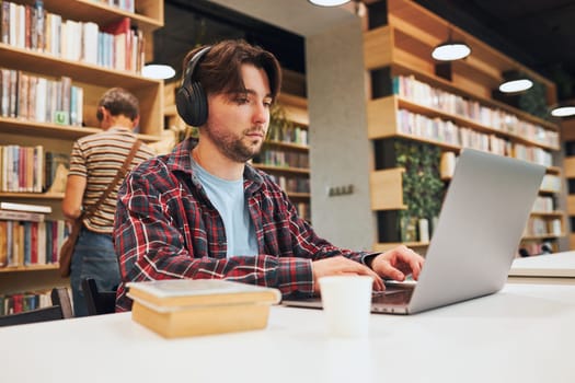 Student learning in university library. Young man preparing for test on laptop. Man listening to online course. Focused student studying for college exams. Using technology