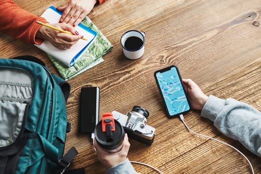 Family planning vacation trip. Girl searching travel destination and routes using navigation map on mobile phone. Woman writing journal and making notes. Charging smartphone with power bank. Using technology while travelling. People sitting at wooden table with camera and backpack during journey. Travel plan concept