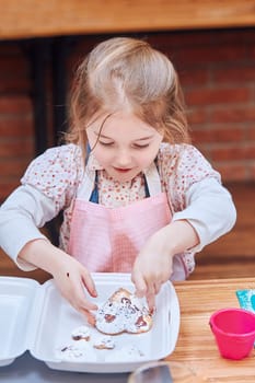 Little girl packing her baked cookies to a box. Kid taking part in baking workshop. Baking classes for children, aspiring little chefs. Learning to cook. Combining and stirring prepared ingredients. Real people, authentic situations