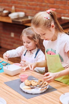 Little girls decorating baked cookies with colorful sprinkle and icing sugar. Kids taking part in baking workshop. Baking classes for children, aspiring little chefs. Learning to cook. Combining and stirring prepared ingredients. Real people, authentic situations