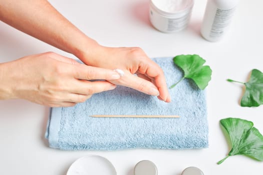 Woman applying  cosmetic moisturizing hand cream. Cosmetic products, green leaves on white table. Spa, manicure, skin care concept. Flat lay, overhead view
